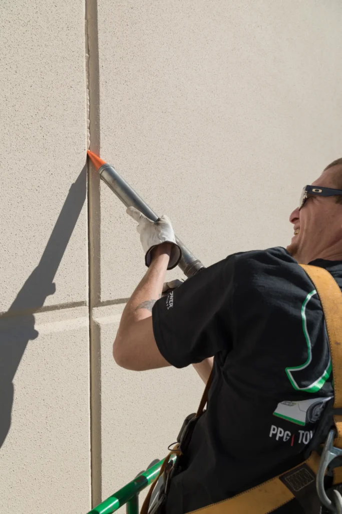 Worker applying sealant to a vertical joint between concrete wall panels on a commercial building.